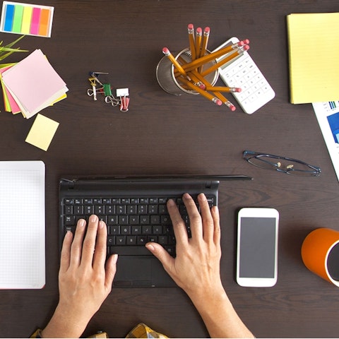 overhead view of hands typing on a wireless keyboard with a cup of pencils, phone, sticky notes and a coffee mug surrounding it