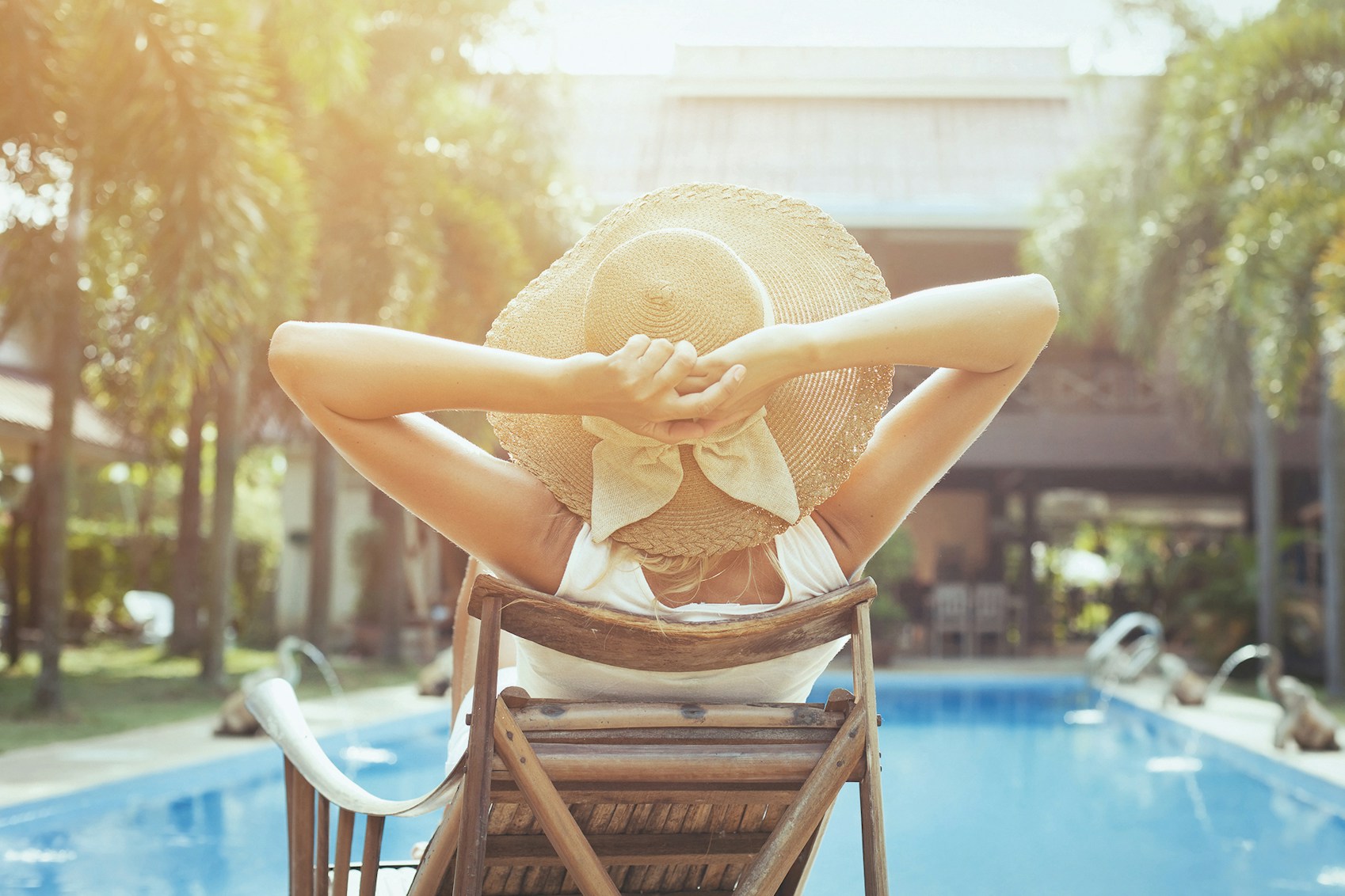view from behind of a lady in a large sun hat sitting in a lounge chair by a pool